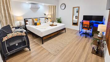A hotel room with a bed, a desk with a computer, a chair, a TV, and a clock on the wall.