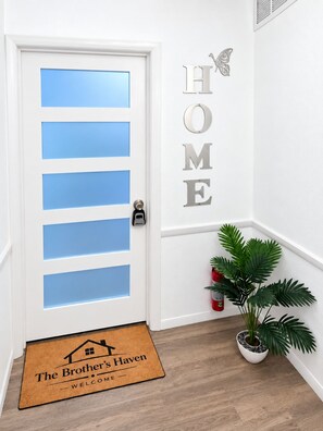 Una porta blanca amb panells blaus, un rètol fixat a la paret que diu 'HOME', un felp de porta amb la inscripció 'The Brother's Haven' i una planta en test.