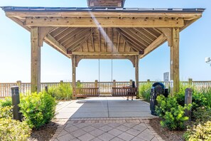 A wooden gazebo with a bench, surrounded by greenery and a paved walkway. - Beach Club Cottage #36 - Dune's Perch (Gulf Shores)