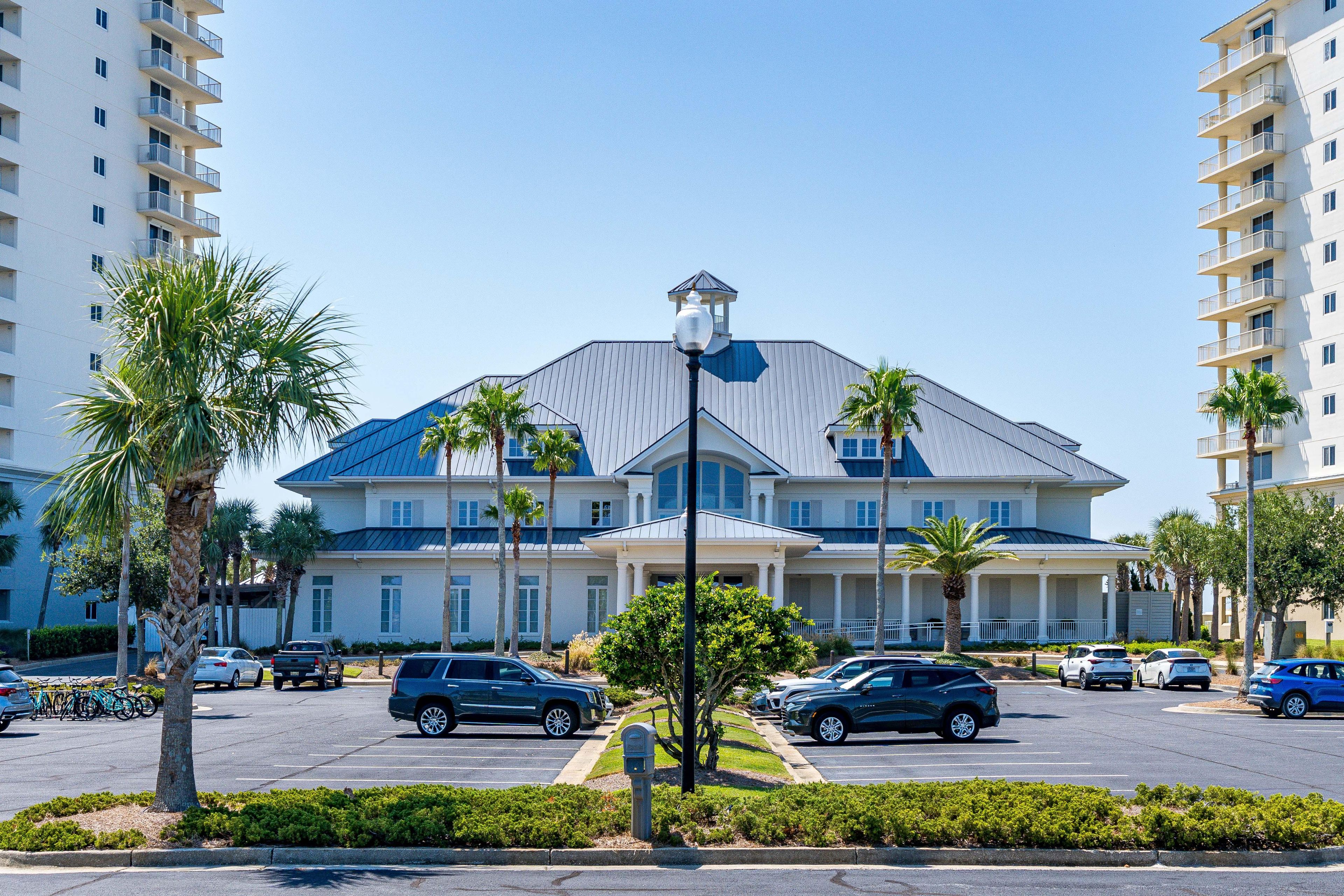 A multi-story hotel with a central entrance, parking lot, and surrounding palm trees.