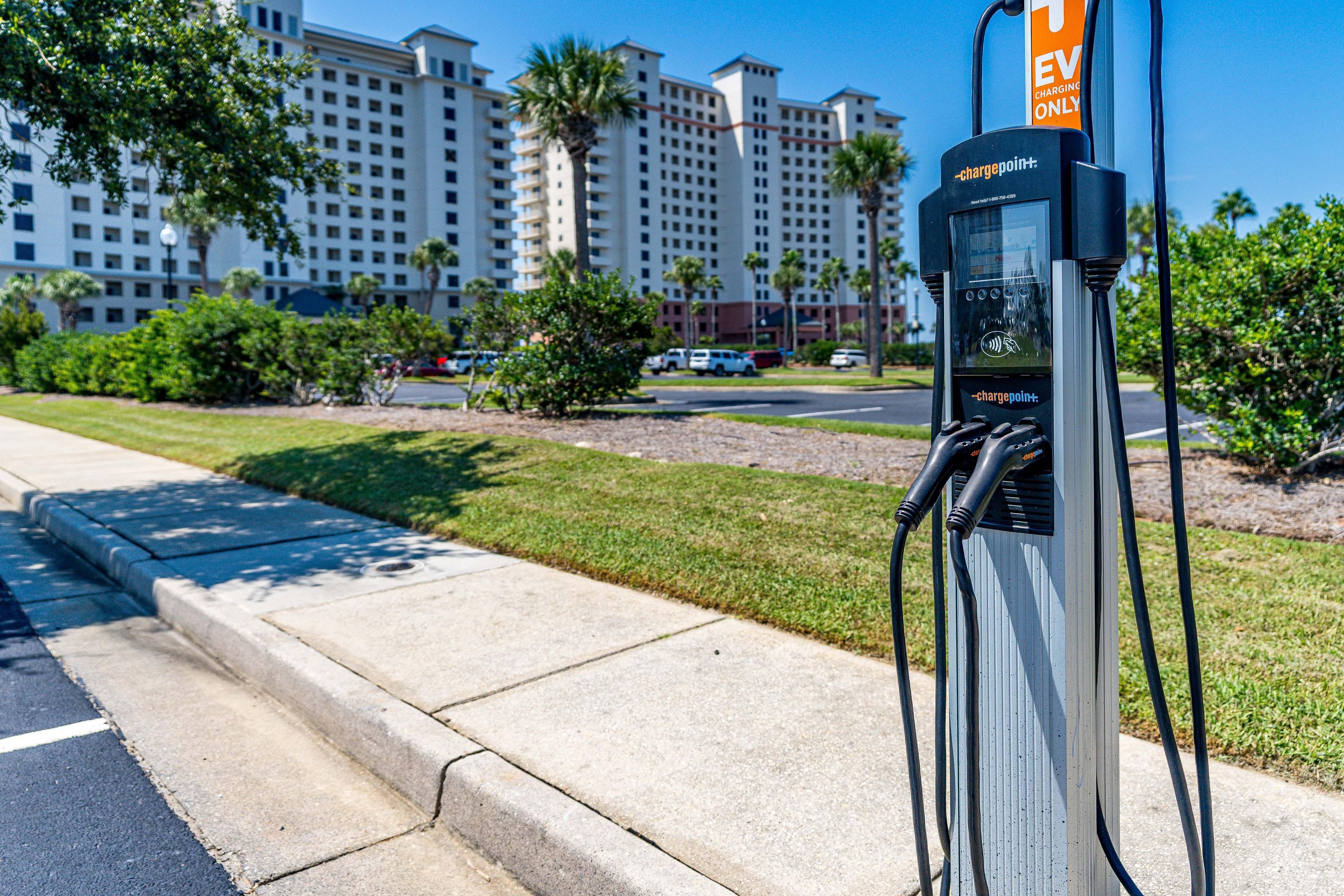 A chargepoint+ electric vehicle charging station on a sidewalk with a road and buildings in the background.