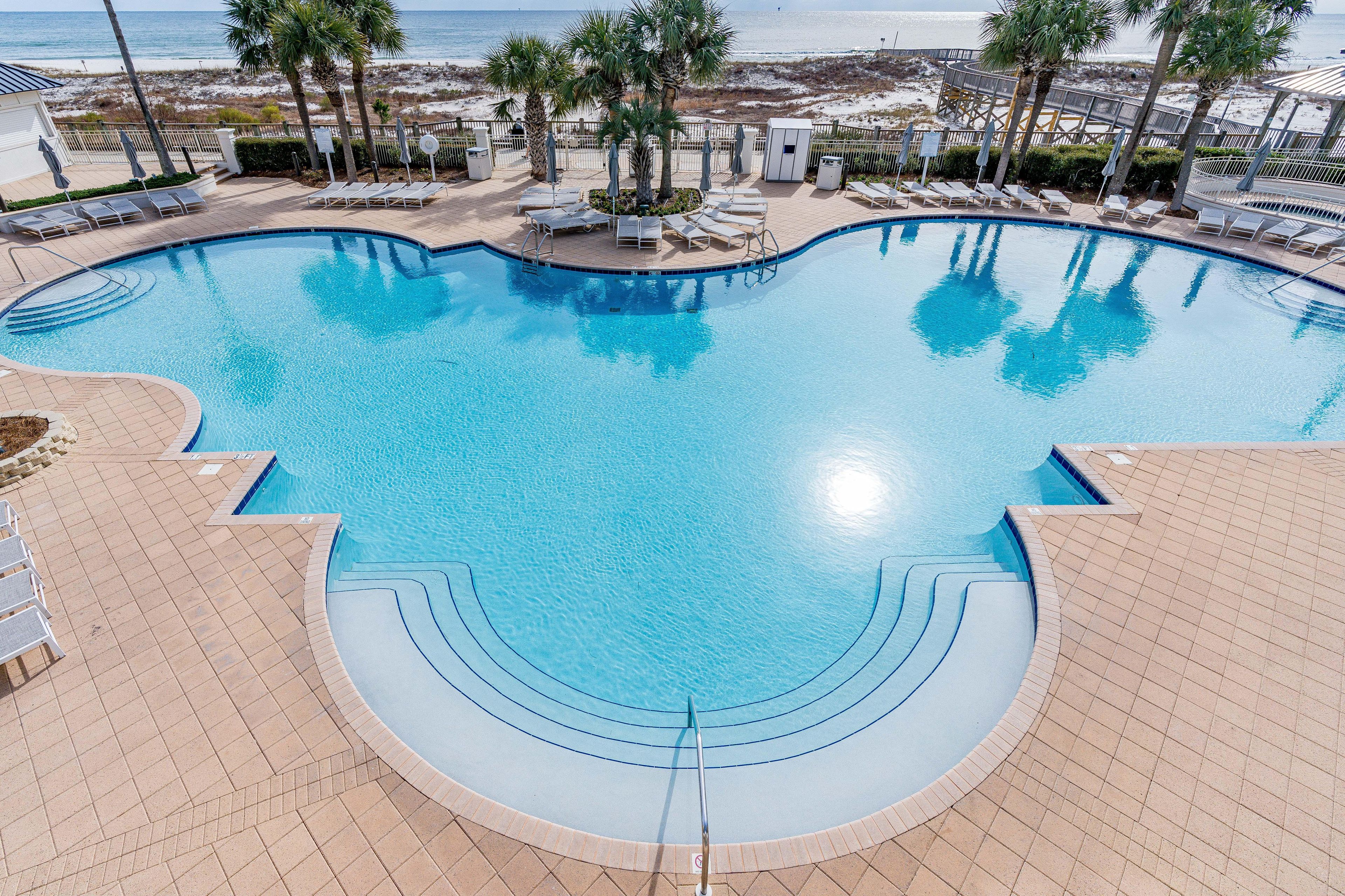 A swimming pool with a curved design, surrounded by lounge chairs and palm trees, with a beach and ocean view in the background.