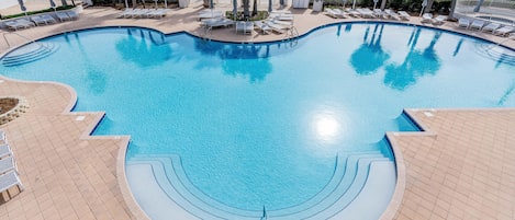 Ein Pool mit geschwungenem Design, umgeben von Liegestühlen und Palmen, mit Strand und Meerblick im Hintergrund.