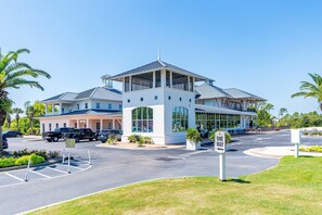 A modern hotel with a central tower, large windows, and a parking area with several vehicles. - Beach Club Cottage #36 - Dune's Perch (Gulf Shores)