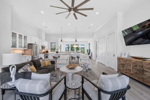 A spacious living room with a modern design, featuring a ceiling fan, a large TV, and a kitchen area in the background. - Beach Club Cottage #36 - Dune's Perch (Gulf Shores)