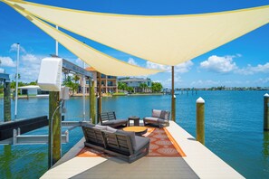 Une terrasse dotée de mobilier d’extérieur, donnant sur une étendue d’eau, avec un bâtiment en arrière-plan et un ciel dégagé.