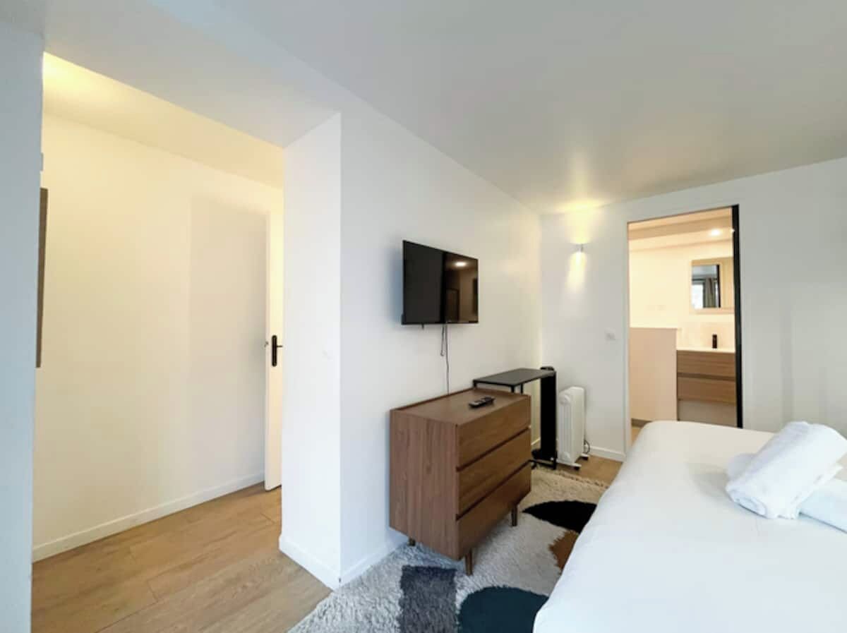 Een moderne slaapkamer met een bed, een dressoir, een televisietoestel en een badkamer die door een open deur zichtbaar is.