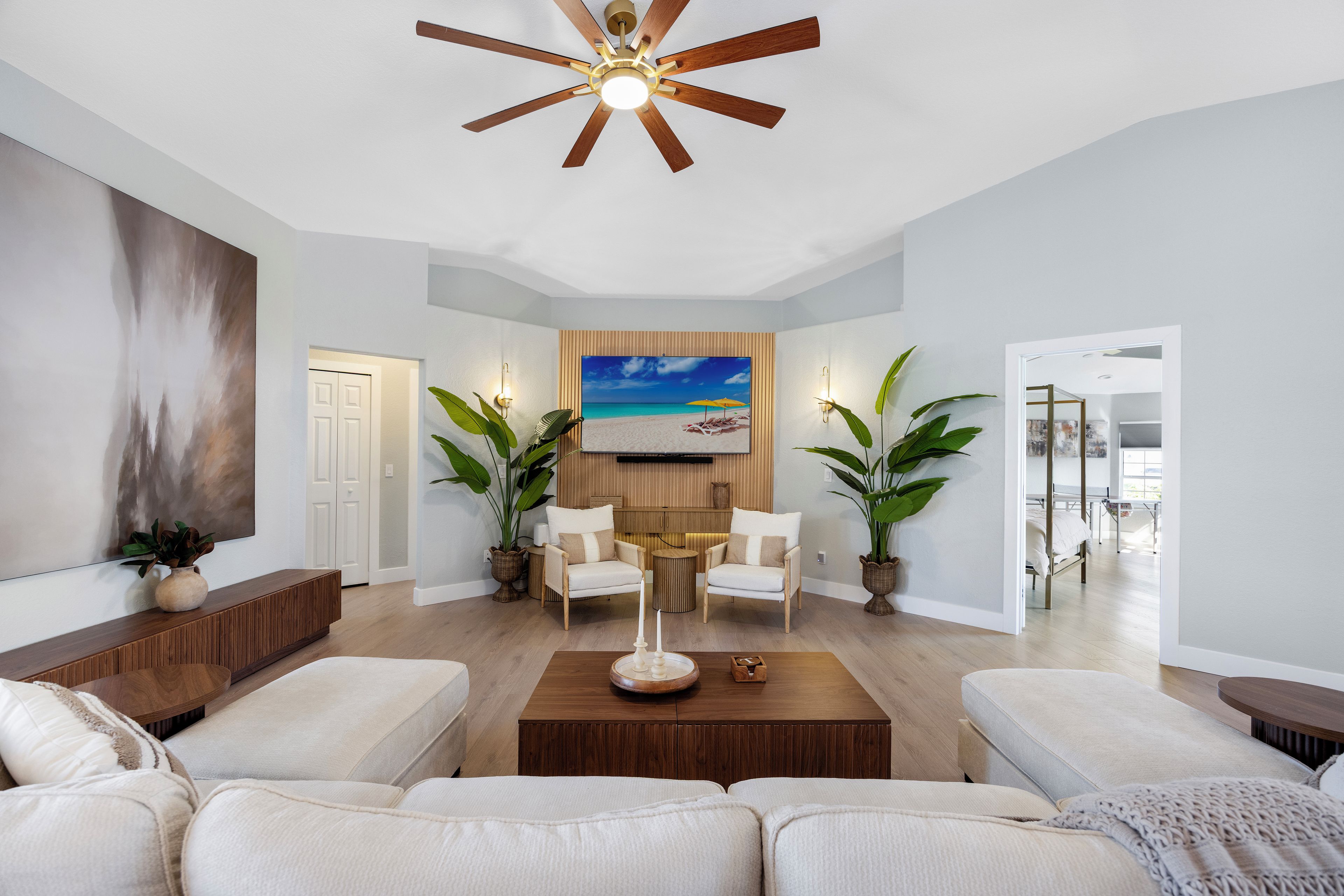 A spacious living room with a ceiling fan, a large painting of a beach scene, two armchairs, a coffee table, and a sofa.