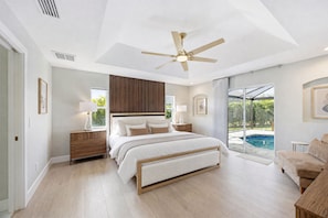 A bedroom with a large bed, a ceiling fan, a nightstand, a lamp, a window with a view of a swimming pool, and a door. - New Luxury 4BR Gulf-Access Canal Villa with Heated Pool, AC, & Central Location (Cape Coral)