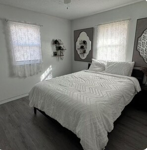 A bedroom with a bed, two windows with sheer curtains, a wall-mounted plant shelf, and a decorative mirror.