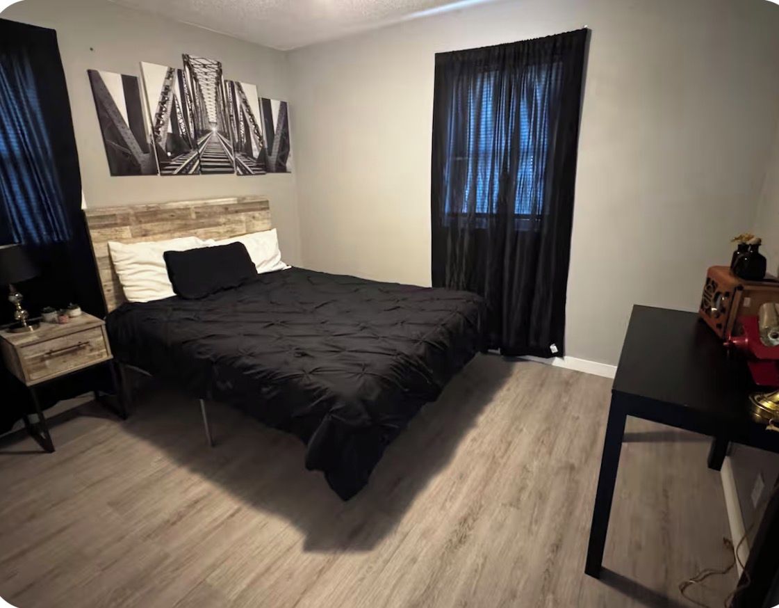 A bedroom with a bed, a nightstand, a dresser, a desk, and a window with curtains.