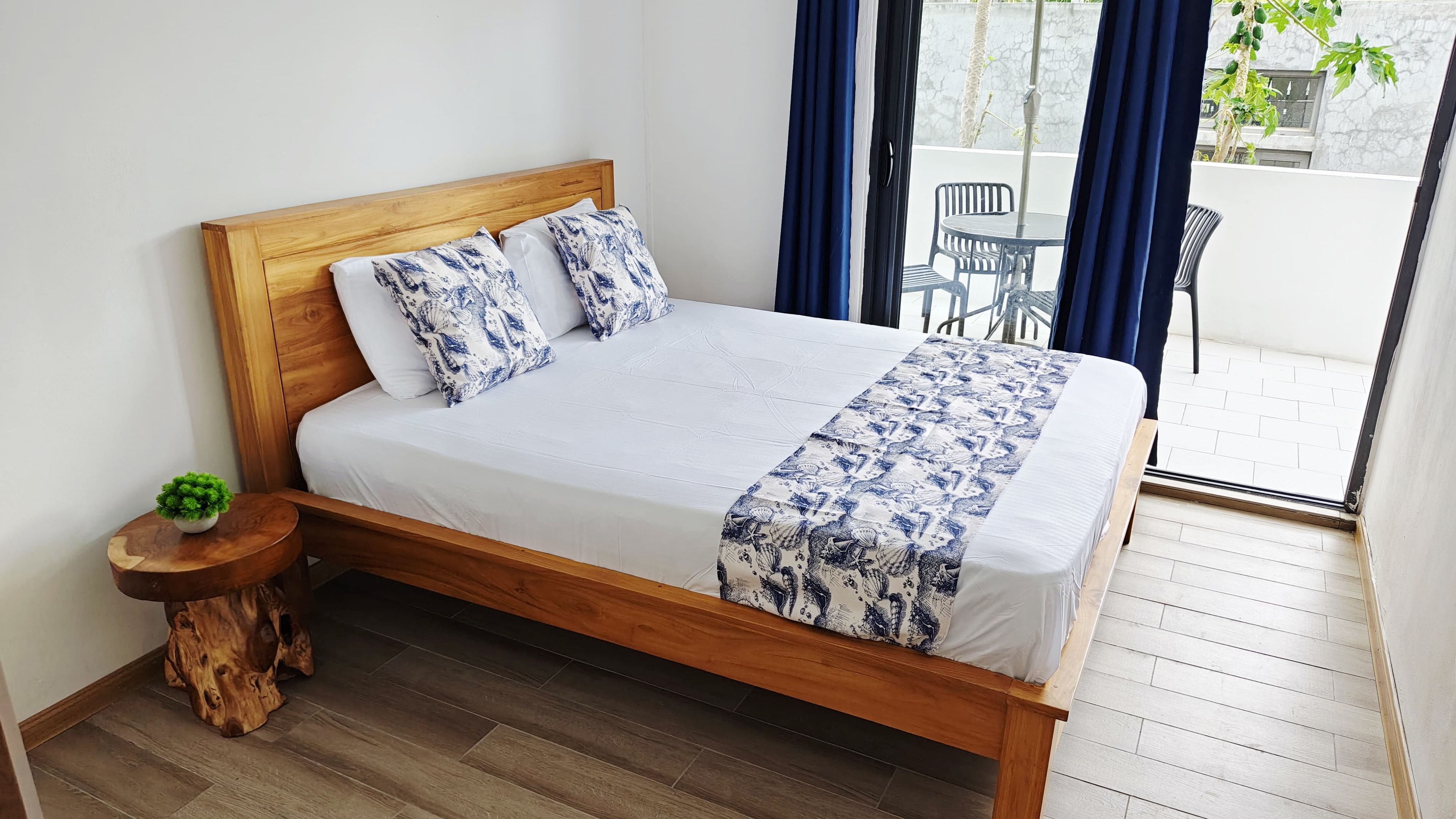 A neatly made bed with a wooden frame, a small bedside table with a plant, and a view of an outdoor area with chairs and a table through a glass door.