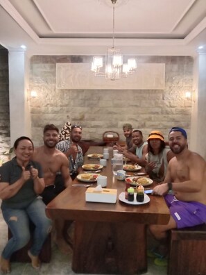 Un grupo de personas sentadas alrededor de una mesa de madera con comida y bebidas, en una habitación con pared de piedra y un candelabro.