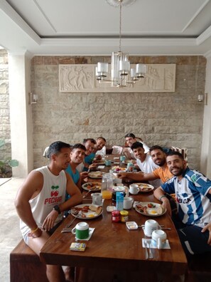 A group of people seated around a wooden dining table with various breakfast items, in a room with a stone wall and a chandelier.