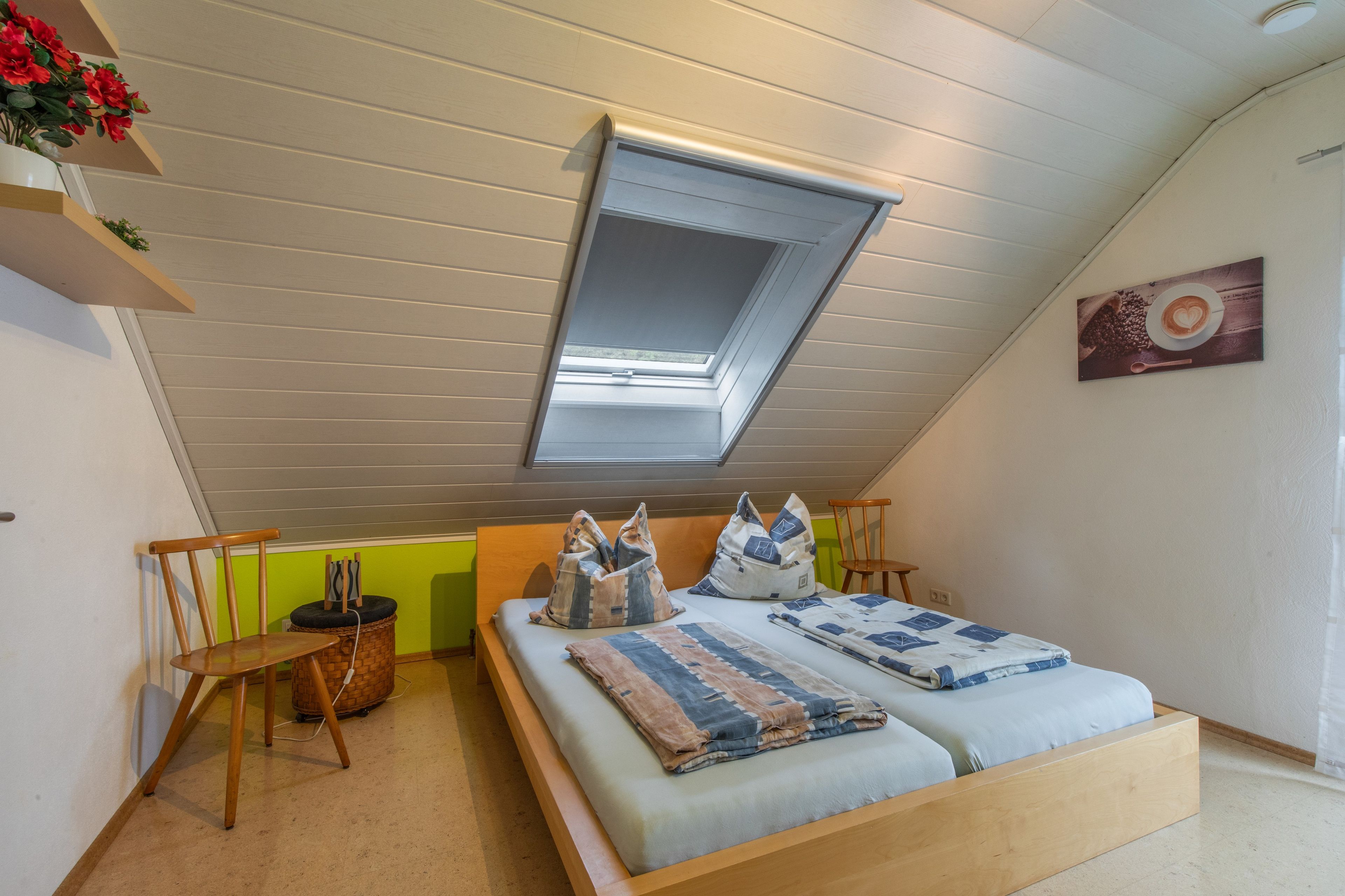 Ein Schlafzimmer mit schrägen Wänden, einem Oberlicht, einem Bett mit Steppdecke, einem Sessel und einem kleinen Tisch.