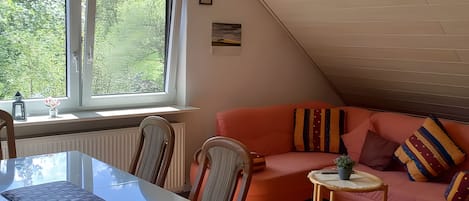 A cozy room with a sloped ceiling, a pink sofa, a small table, and a window with a view of greenery.