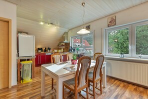 A modern kitchen with wooden flooring, a dining table with chairs, and a view of a red-roofed house through large windows.