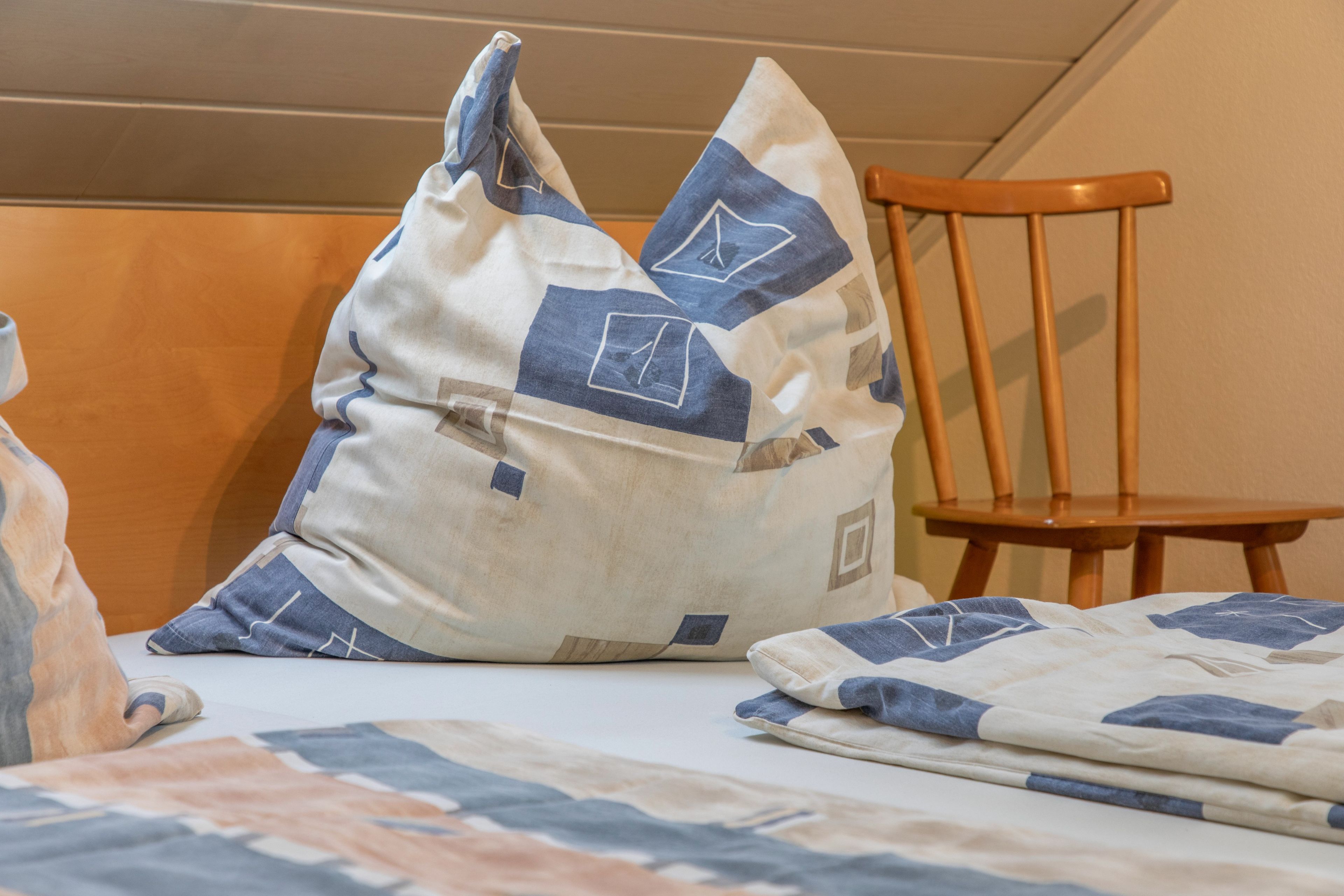 A bed with a blue and white patterned bedspread and pillows, a wooden chair, and a wall with a horizontal panel design.