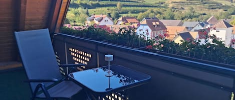 Varanda com cadeira e mesa, com vista para uma colina com casas e vinhedos.