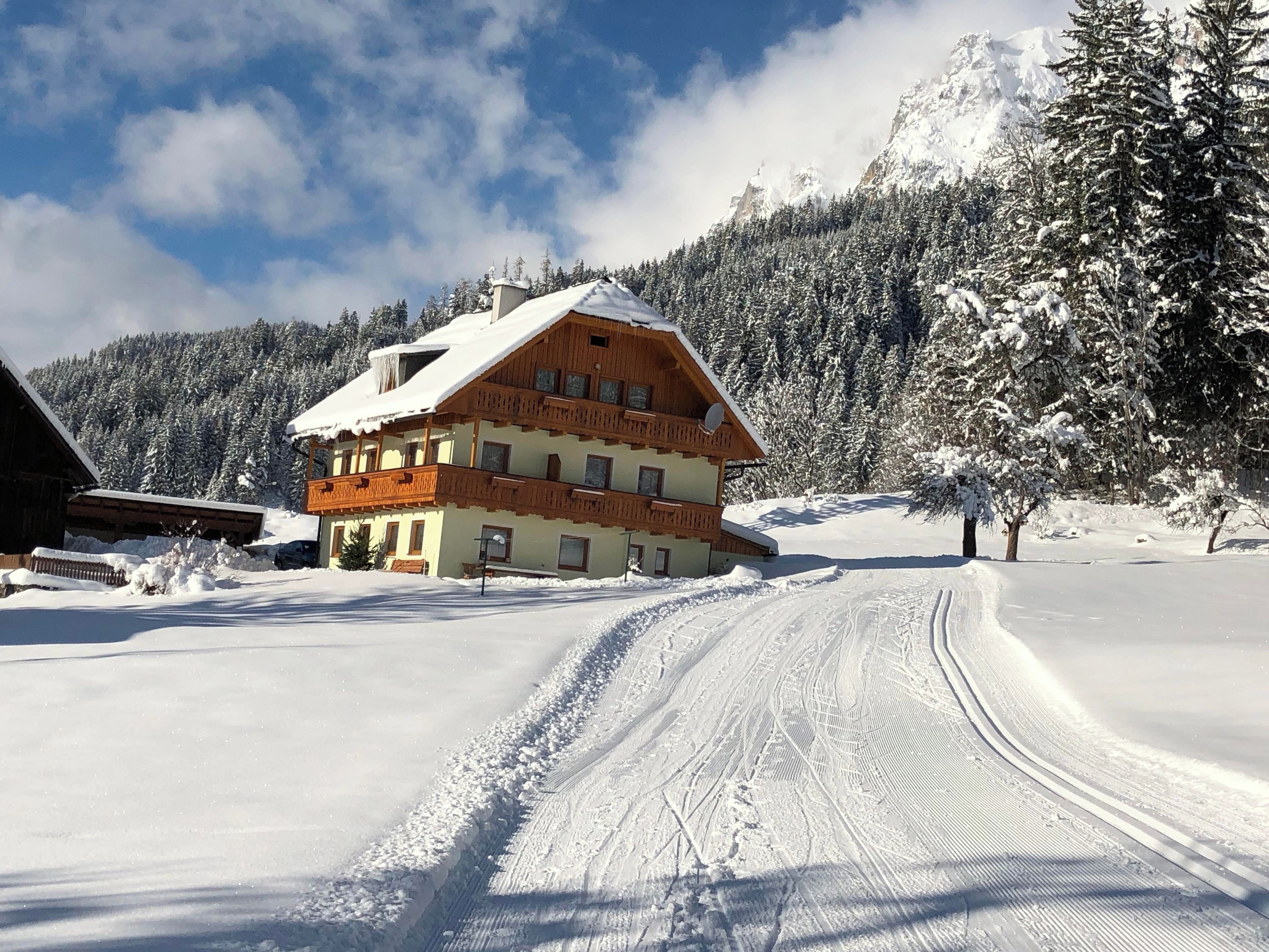 Een meerlaags houten chalet met een met sneeuw bedekte kap, gelegen in een besneeerd landschap met een bebost berglandschap op de achtergrond.