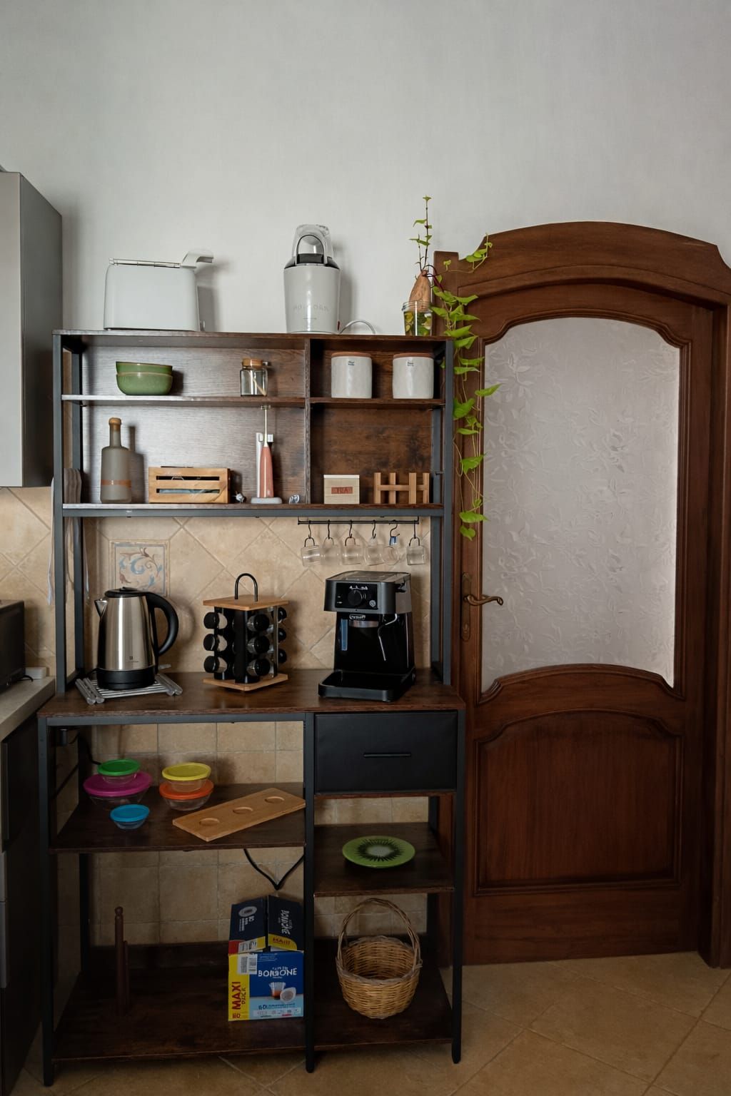 A kitchen with a coffee machine, shelves with jars, a kettle, and a wooden cabinet.