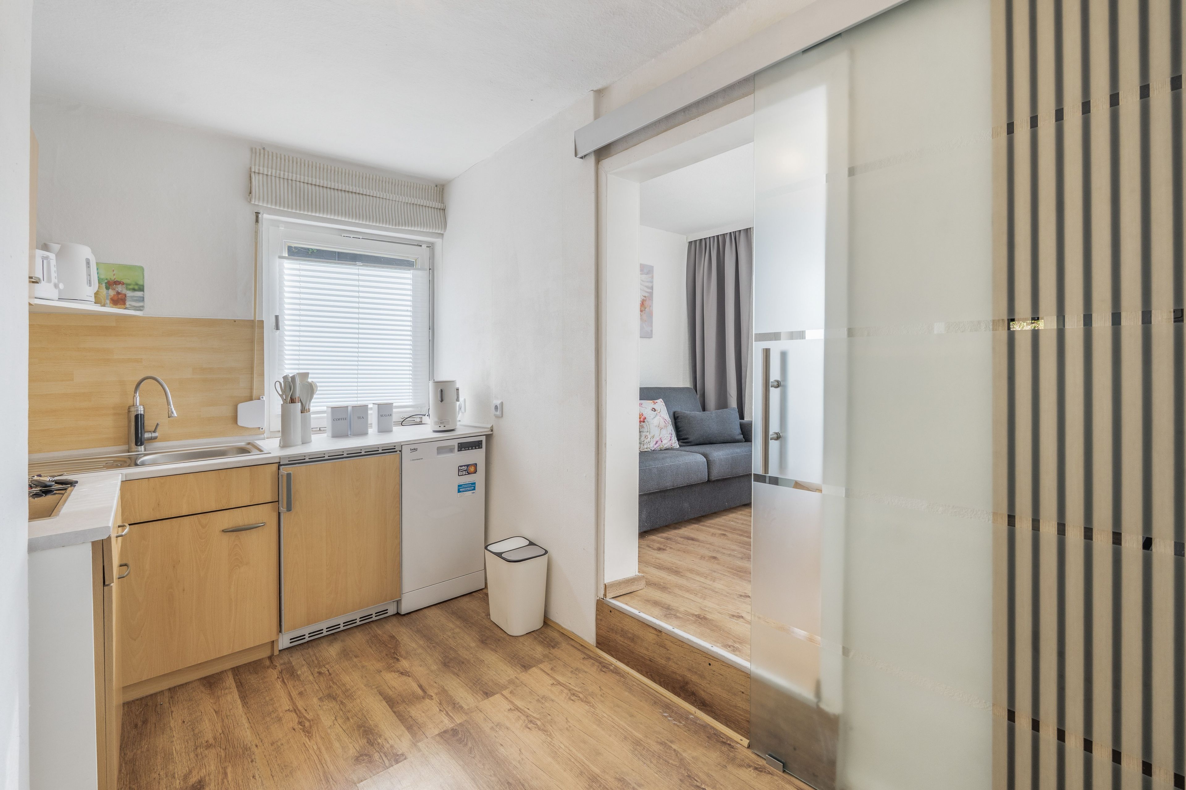 A compact kitchen with a built-in dishwasher, a sink, and a window with blinds.