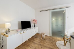 Un saló modern amb un televisor pantalla plana sobre un moble blanc, un tamboret de fusta i una porta de vidre que dona a un balcó.