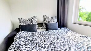 Een slaapkamer met een bed, twee kussens, een raam met gordijnen en uitzicht op groen.