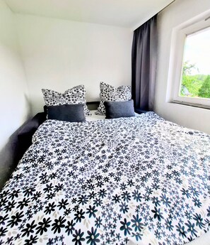 Une chambre à coucher avec un lit, deux oreillers, une fenêtre avec des rideaux et une vue sur la verdure à l’extérieur.