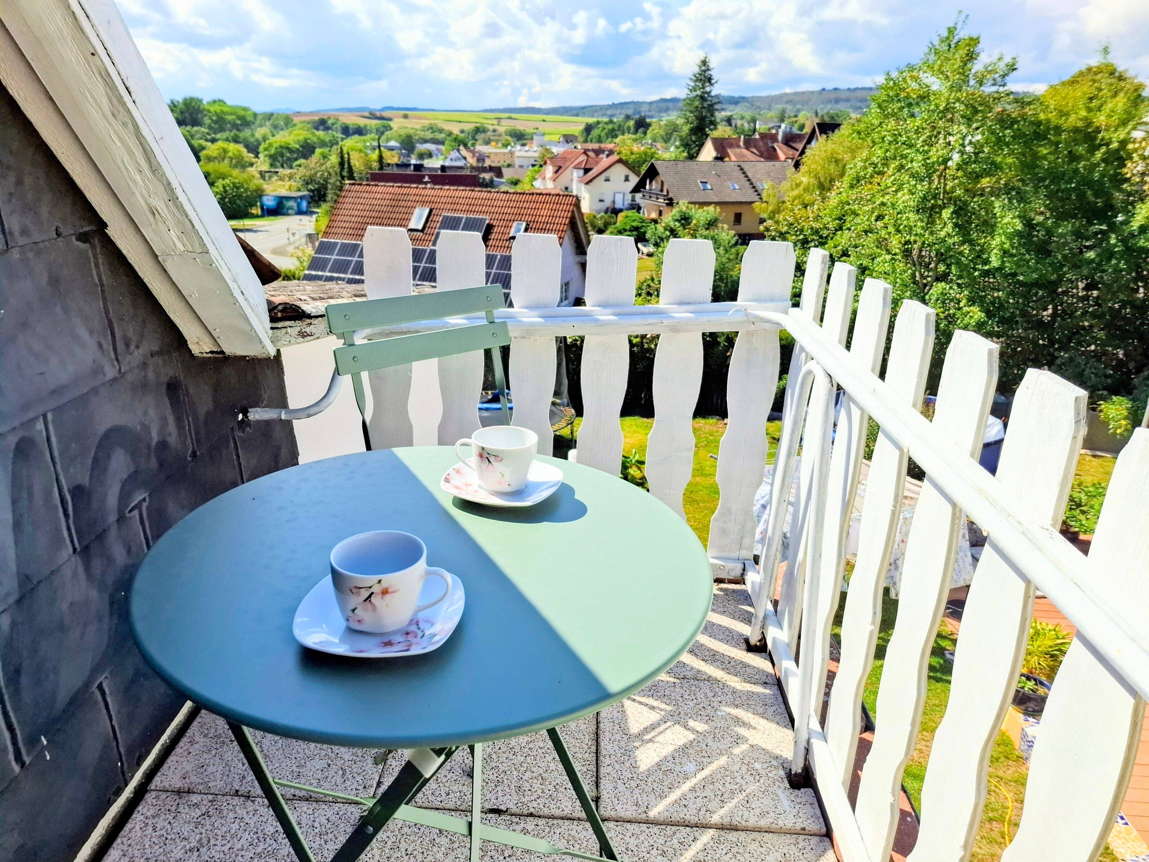 Un balcon avec une table et deux tasses, donnant sur un village avec des maisons et de la verdure.