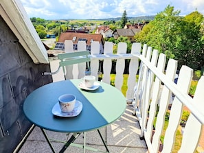 Un balcon avec une table et deux tasses, donnant sur un village avec des maisons et de la verdure.