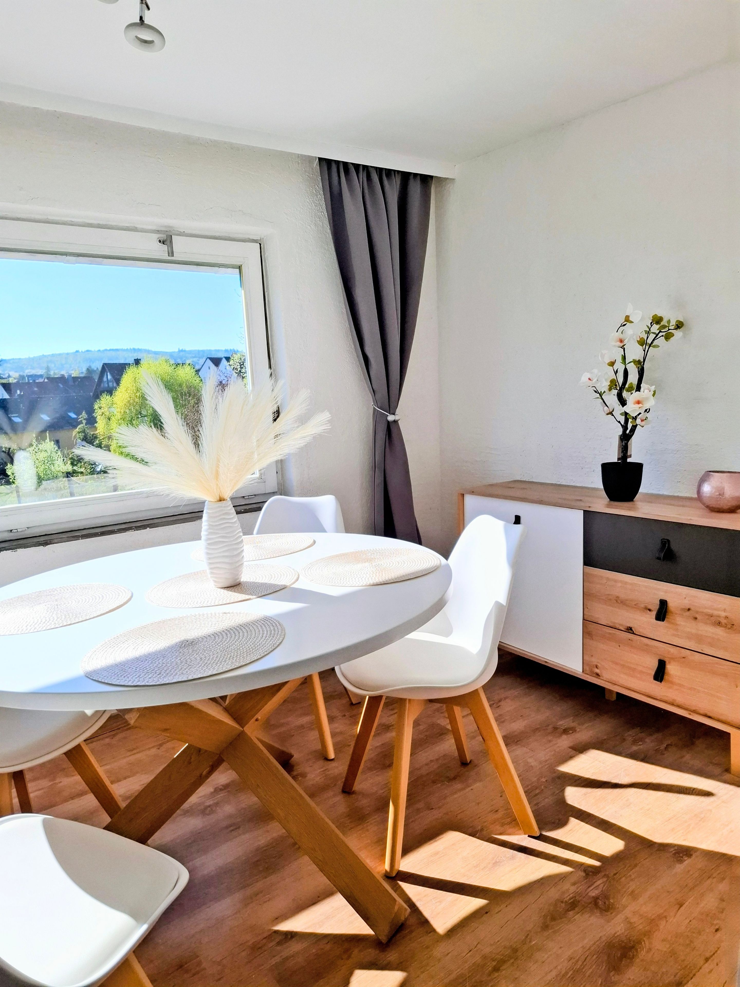 Ein moderner Essbereich mit einem runden Tisch, weißen Stühlen, einer Vase mit Trockenpflanzen und Blick auf Grün.