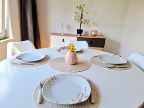 Een ronde eettafel met witte borden en bestek, een geweven tafelmat en een kleine decoratieve vaas met gele bloemen.