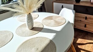 Een eetgedeelte met een ronde tafel, geweven placemats, een vaas met droogbloemen en een potplant op een houten kast.