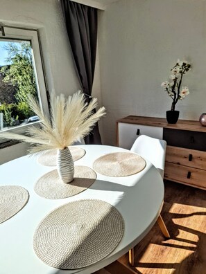 Un coin repas avec une table ronde, des sets de table en fibres naturelles, un vase avec des fleurs séchées et une plante en pot posée sur un meuble en bois.