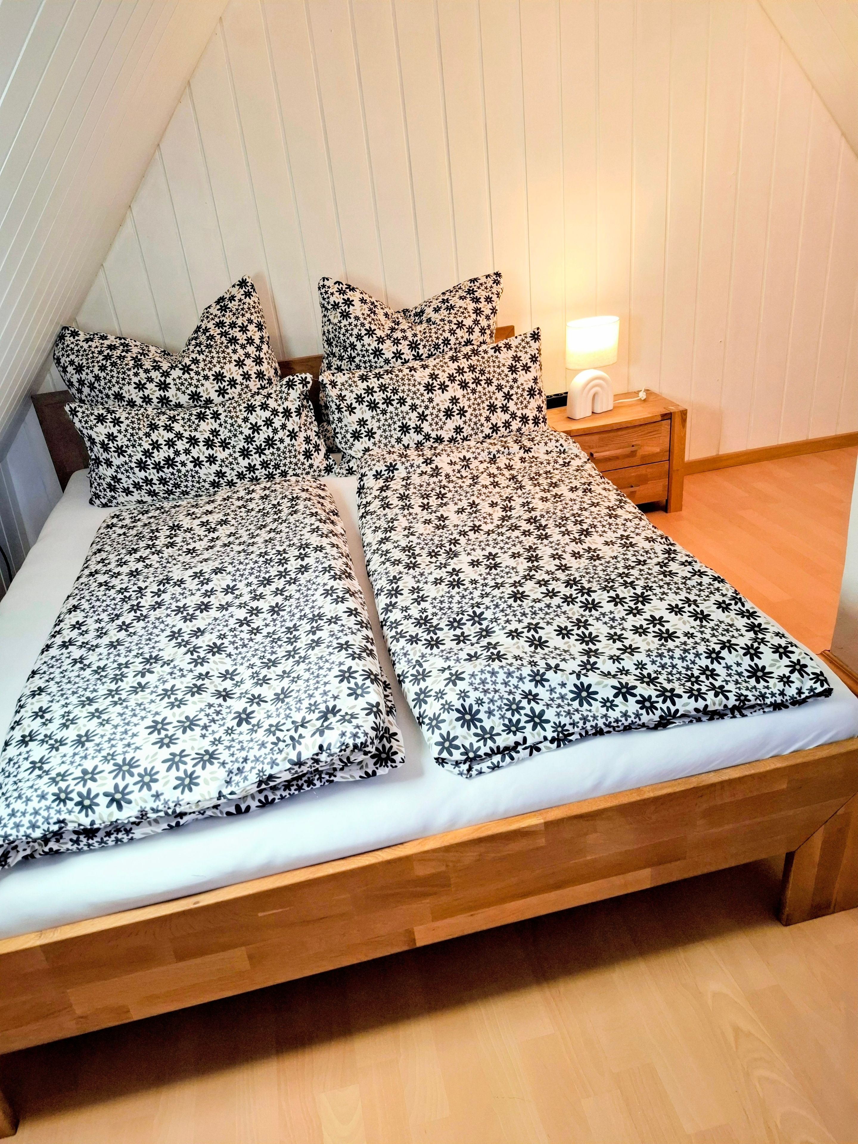 Ein Bett mit blumigem Bettbezug und Kissen, ein Holz-Kopfteil, ein Nachttisch mit Lampe und ein Holzboden.