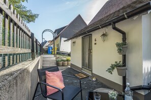 Een smalle buitenpassage met een stoel en een tafeltje, een gebouw met een pannendak en een metalen hek aan de zijkant.