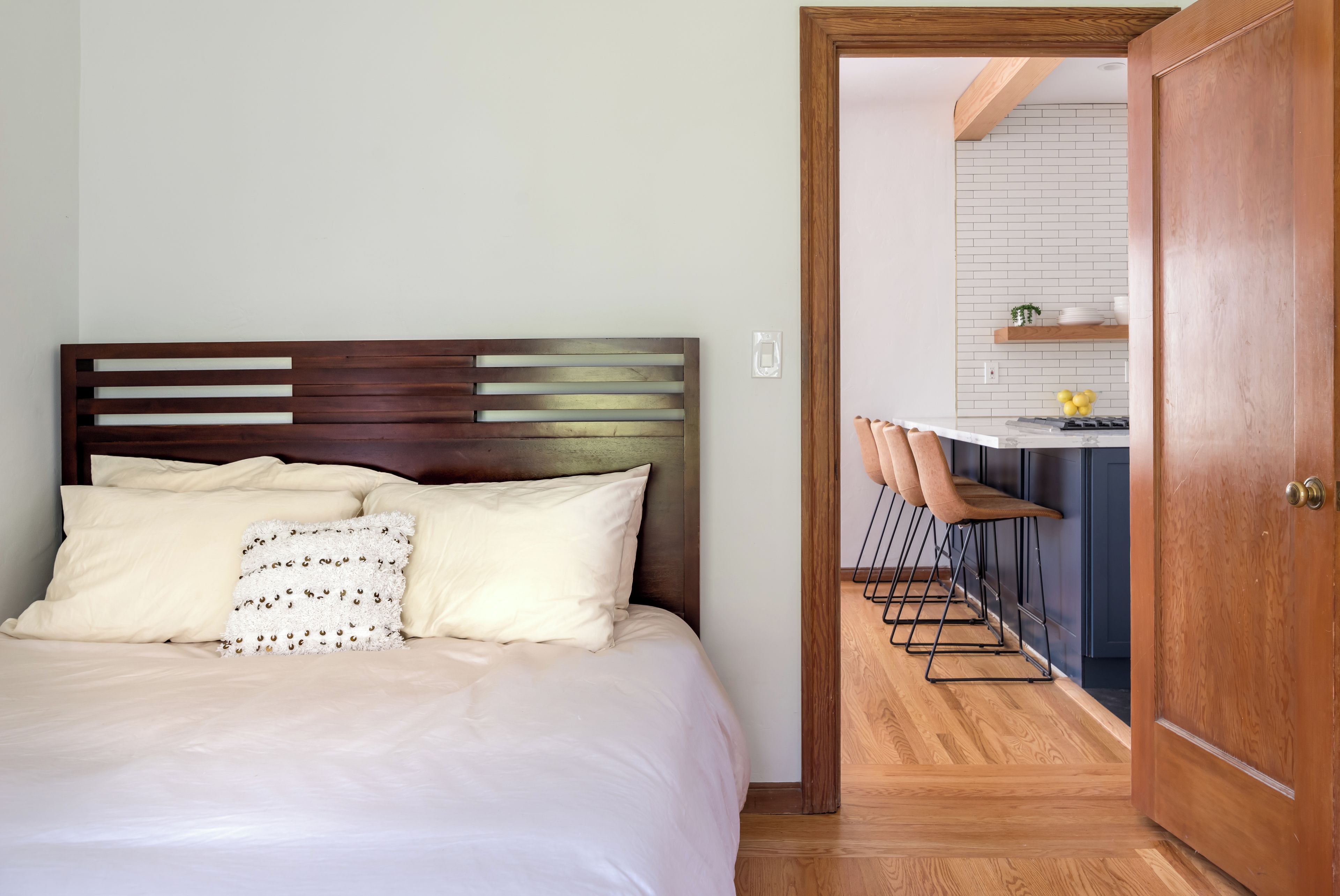 Une chambre à coucher avec une tête de lit en bois, un lit avec une literie blanche et une vue sur un coin cuisine équipé d’un bar et de tabourets.