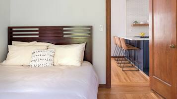 Un dormitorio con cabecera de madera, cama con ropa de cama blanca y vista hacia una cocina con barra y taburetes.