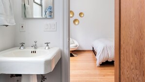A bathroom with a white sink, mirror, and three wall-mounted lights. There is a wooden door leading to a bedroom with a bed and a small round table. - Stunning Two Bedroom Mediterranean Home  (Oakland)
