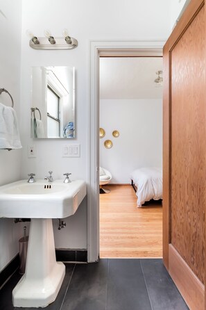 Un baño con un lavamanos blanco, un espejo y tres luces empotradas en la pared. Hay una puerta de madera que da a un dormitorio con una cama y una mesita redonda.