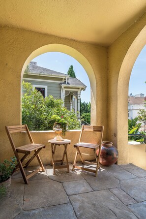 Un patio con dos sillas de madera y una mesa pequeña, una olla grande de cobre, y una vista de una casa a través de un arco.