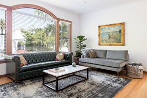 Una sala de estar con un sofá de cuero verde, un sofá gris, una mesa de centro, un cuadro y una ventana con persianas.