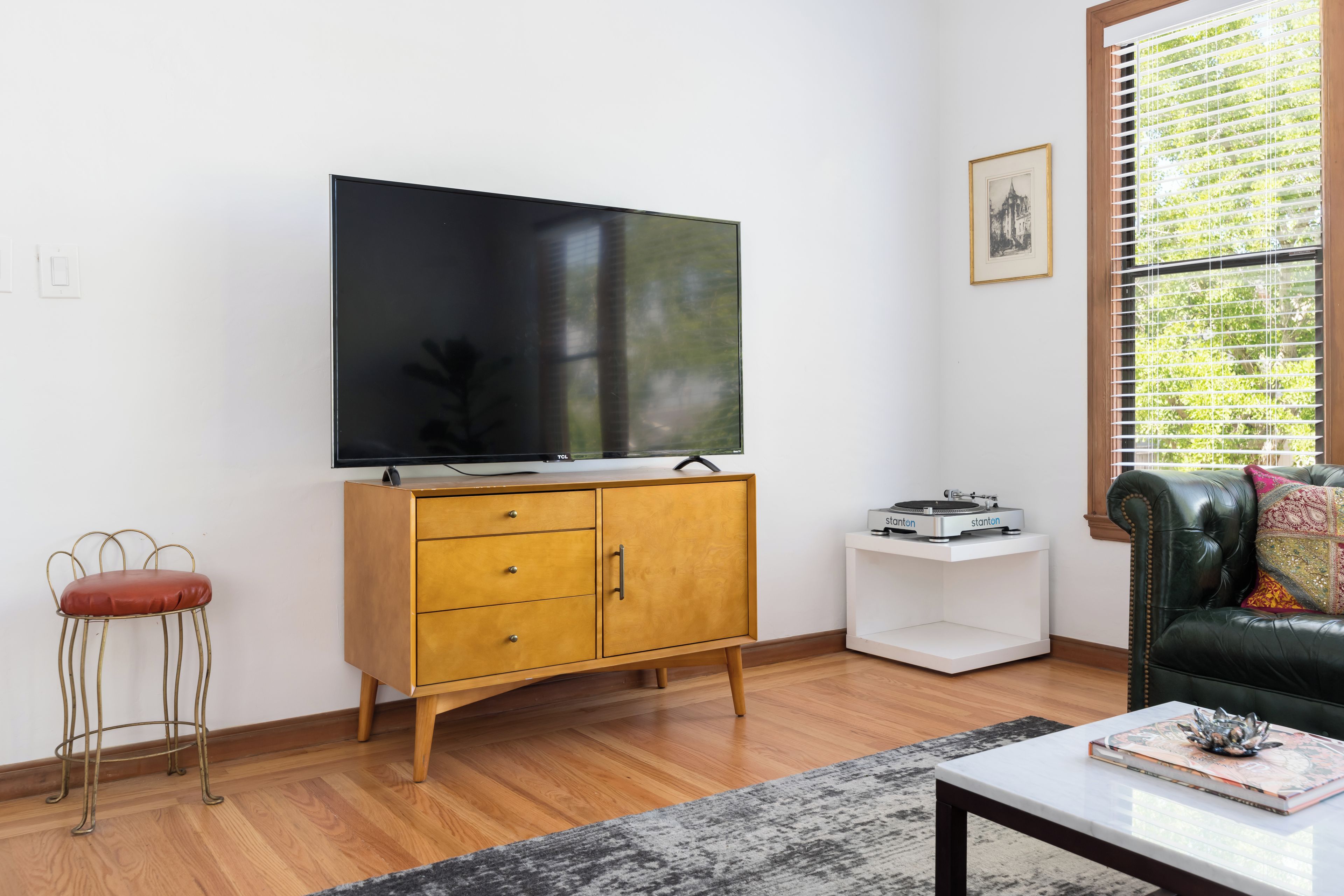 Una sala de estar con televisor de pantalla plana, mueble de madera, pequeña mesita auxiliar blanca y un sofá.