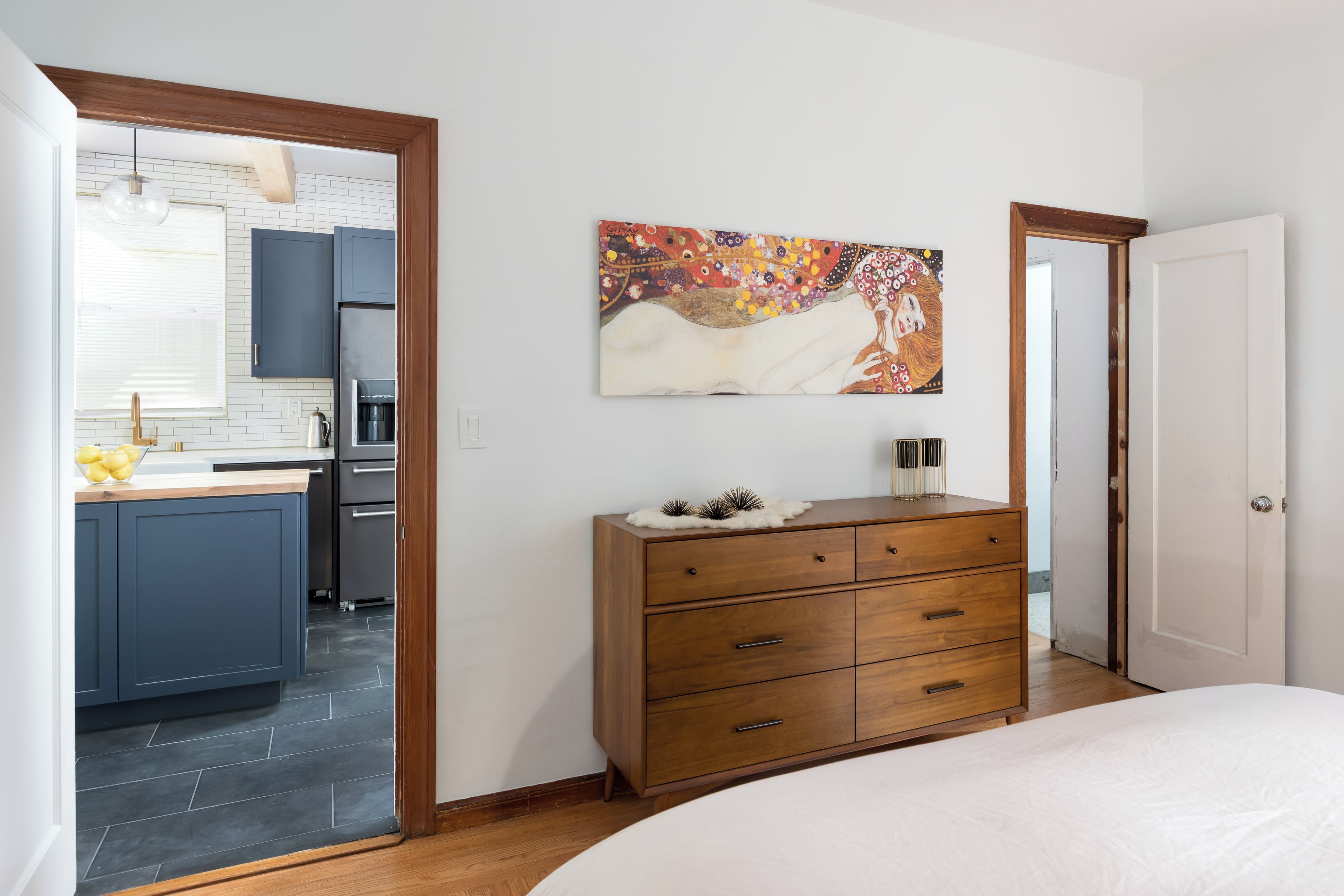Ein Schlafzimmer mit einem Holzkasten, einem Bett und einer durch eine offene Tür einsehbarer Küche.