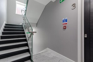 A staircase with glass railings, a fire action sign, and a door with a number plate. - Aldgate East by Excel Apartments (London)