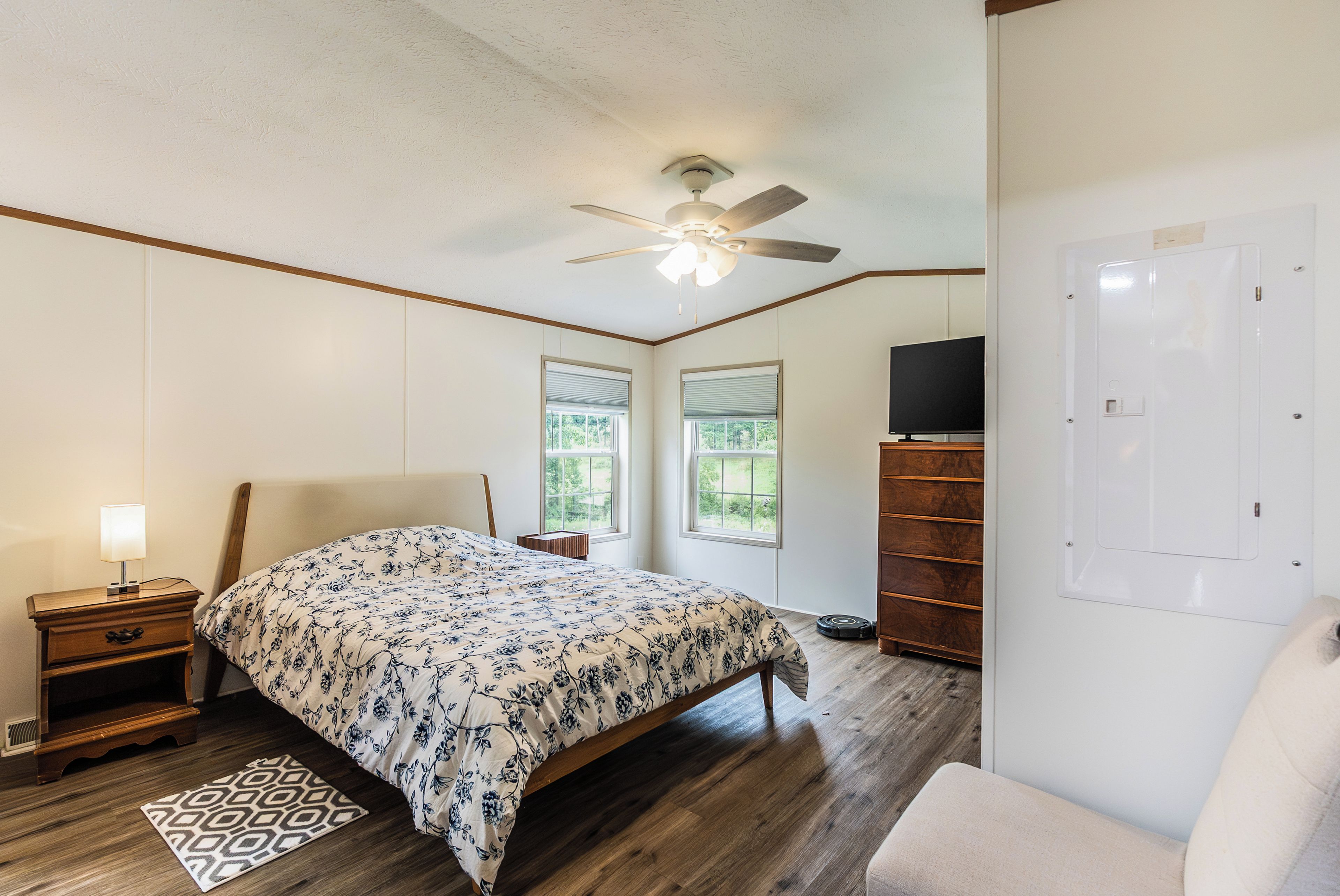 A bedroom with a bed, nightstand, lamp, ceiling fan, windows, and a TV.