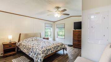 A bedroom with a bed, nightstand, lamp, ceiling fan, windows, and a TV.