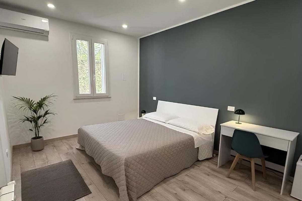 A bedroom with a bed, bedside table, desk, chair, potted plant, and a window.