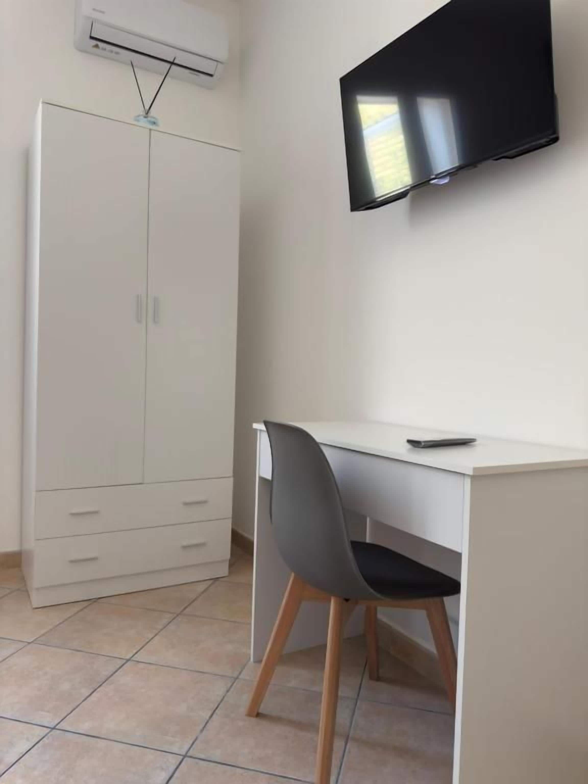 Una habitación con un escritorio blanco, una silla gris y un televisor de pantalla plana montado en la pared.